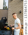 Grilling food outdoors in a backyard during a sunny day with a man preparing meat and vegetables on a barbecue 137864753