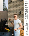 Man grilling meat and vegetables outside near a modern building on a sunny day, enjoying time with friends and family 137864754