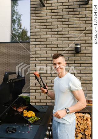 Man grilling meat and vegetables outside near a modern building on a sunny day, enjoying time with friends and family Man grilling meat and vegetables outside near a modern building on a sunny day, enjoying time with friends and family 137864754