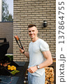 Man grilling meat and vegetables outside near a modern building on a sunny day, enjoying time with friends and family 137864755