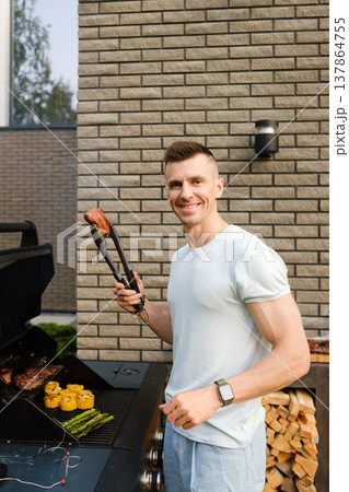 Man grilling meat and vegetables outside near a modern building on a sunny day, enjoying time with friends and family 137864755