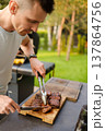 Man slices grilled meat outside in backyard during a sunny day with green trees around and summer vibe in the air 137864756