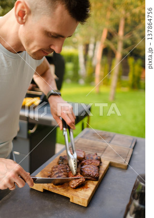 Man slices grilled meat outside in backyard during a sunny day with green trees around and summer vibe in the air Man slices grilled meat outside in backyard during a sunny day with green trees around and summer vibe in the air 137864756