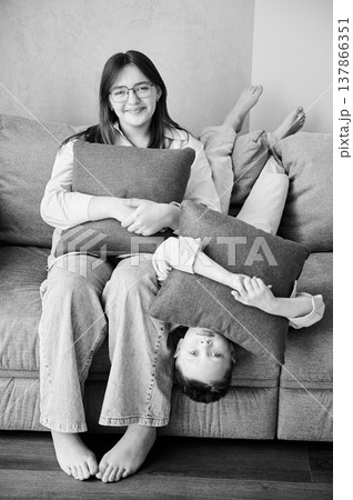 Young siblings relaxing on a couch. Happy sister teenage girl with glasses and brother laying upside down on couch. Smiling kids hugging pillows. Black and white image. 137866351
