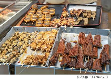 Assorted Japanese grilled seafood and dango skewers displayed at a market stall. Includes unagi eel, oysters, scallops, and octopus with savory glazes on metal trays for authentic street food dining. Assorted Japanese grilled seafood and dango skewers displayed at a market stall. Includes unagi eel, oysters, scallops, and octopus with savory glazes on metal trays for authentic street food dining. 137866784