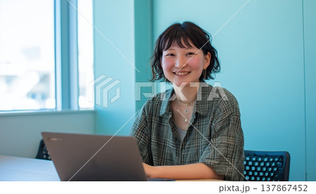 日中の青い壁が特徴的なオフィスでノートパソコンに向かいながら明るく微笑んでいる若い女性 日中の青い壁が特徴的なオフィスでノートパソコンに向かいながら明るく微笑んでいる若い女性 137867452
