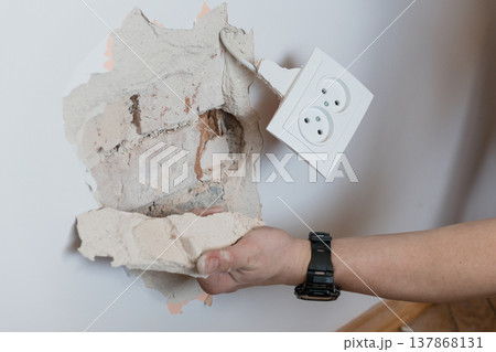 Damaged Drywall Wall With Broken Electrical Outlet Being Held By Hand Showing Exposed Plaster And Wiring In White Room Interior Scene 137868131
