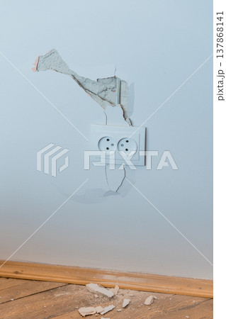 Damaged White Wall With Cracked Plaster Around Electrical Outlet Showing Exposed Concrete And Broken Pieces On Wooden Floor In Light Blue Room Damaged White Wall With Cracked Plaster Around Electrical Outlet Showing Exposed Concrete And Broken Pieces On Wooden Floor In Light Blue Room 137868141