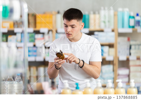 Young male buyer choosing medicinal cough liquid syrup standing between shelves at pharmacy Young male buyer choosing medicinal cough liquid syrup standing between shelves at pharmacy 137868885