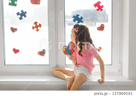 A lonely sad girl is sitting on the windowsill by the window, decorated with pieces of multicolored puzzles and hearts with card in bunches. Decorated room for Autism Day 137869581