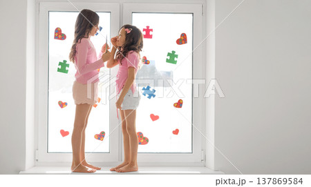 The girls are standing on the windowsill, jointly decorating the window glass with puzzle pieces and hearts. A banner in support of people with autism spectrum disorder. The girls are standing on the windowsill, jointly decorating the window glass with puzzle pieces and hearts. A banner in support of people with autism spectrum disorder. 137869584