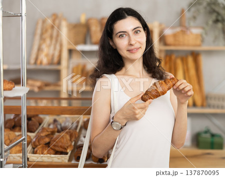 Young female customer stands with croissant in hands near window of bakery. 137870095