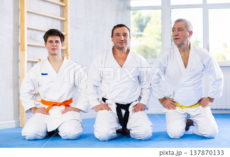 Sports team of different ages in white kimonos sit on their knees on the tatami in gym Sports team of different ages in white kimonos sit on their knees on the tatami in gym 137870133
