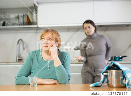 Sad mature woman with soup in pan on table listens unpleasant sayings of adult daughter. Sad mature woman with soup in pan on table listens unpleasant sayings of adult daughter. 137870135