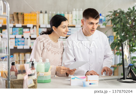 Young professional male parmacist working computer and helping young woman with prescription in hand look for medicine in drugstore Young professional male parmacist working computer and helping young woman with prescription in hand look for medicine in drugstore 137870204