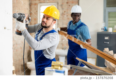 Room renovation - worker drills with puncher, another carries wooden planks Room renovation - worker drills with puncher, another carries wooden planks 137870208