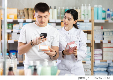 Handsome young man choosing box with pills in pharmacy, scan QR code on label and professional pharmacist woman helping and offering supplements at drugstore Handsome young man choosing box with pills in pharmacy, scan QR code on label and professional pharmacist woman helping and offering supplements at drugstore 137870238