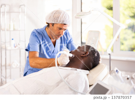 Male cosmetologist performs skin cleansing procedure for African man client with beauty machine Male cosmetologist performs skin cleansing procedure for African man client with beauty machine 137870357