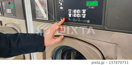 A man presses a button on the control panel of a washing machine at a self-service laundromat. Concepts of everyday life, automation, household services, and the use of modern household appliances A man presses a button on the control panel of a washing machine at a self-service laundromat. Concepts of everyday life, automation, household services, and the use of modern household appliances 137870731
