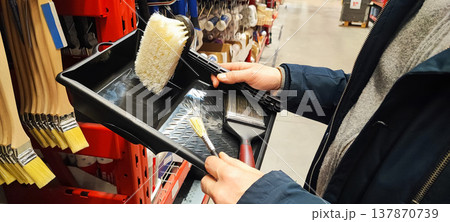 A customer holds a paint tray and brushes while shopping in a hardware store aisle. Concepts of home improvement, DIY renovation, consumer choice, and retail shopping behavior. A customer holds a paint tray and brushes while shopping in a hardware store aisle. Concepts of home improvement, DIY renovation, consumer choice, and retail shopping behavior. 137870739