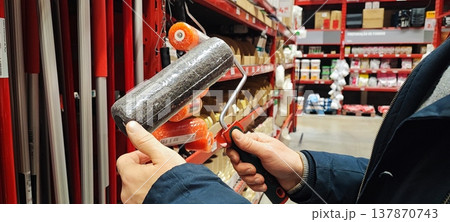 A customer holds a paint tray and brushes while shopping in a hardware store aisle. Concepts of home improvement, DIY renovation, consumer choice, and retail shopping behavior. 137870743