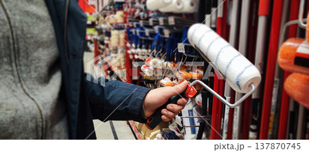 A customer holds a paint tray and brushes while shopping in a hardware store aisle. Concepts of home improvement, DIY renovation, consumer choice, and retail shopping behavior. 137870745