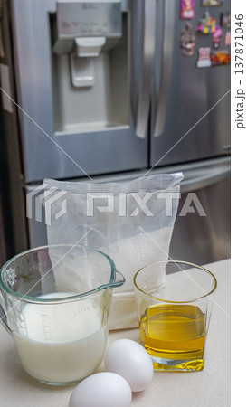 Ingredients for baking displayed on kitchen countertop with eggs, milk, oil, and flour in measuring containers near a refrigerator 137871046