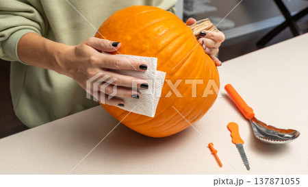 Person cleaning a bright orange pumpkin with a paper towel, surrounded by carving tools on a light countertop in a cozy indoor setting Person cleaning a bright orange pumpkin with a paper towel, surrounded by carving tools on a light countertop in a cozy indoor setting 137871055