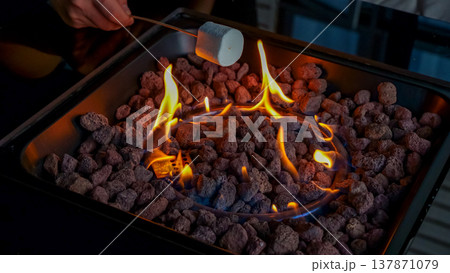 Person roasting marshmallow over flames in a fire pit filled with decorative stones, creating a cozy outdoor atmosphere Person roasting marshmallow over flames in a fire pit filled with decorative stones, creating a cozy outdoor atmosphere 137871079