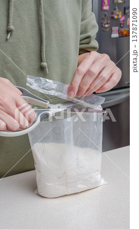 Person using scissors to open a plastic bag containing white powder on a kitchen countertop with a refrigerator in the background 137871090