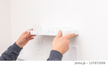 Mans hands holding white rectangular shelf plank against white wall near installed black bracket. Home improvement, positioning shelf board, DIY assembly preparation, fitting stage. 137871099