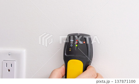 Mans hand using yellow and black stud finder on white wall beside electrical socket. Home improvement, stud detection, DIY task, tool in use. Mans hand using yellow and black stud finder on white wall beside electrical socket. Home improvement, stud detection, DIY task, tool in use. 137871100