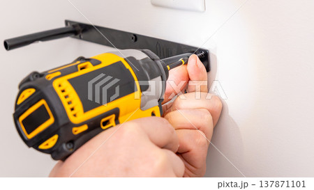 Mans hands using yellow cordless drill to drive screw into white wall bracket attachment. Home improvement, mounting hardware, DIY installation, power tool action. Mans hands using yellow cordless drill to drive screw into white wall bracket attachment. Home improvement, mounting hardware, DIY installation, power tool action. 137871101