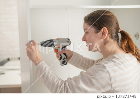 A Caucasian woman uses a screwdriver to tighten a screw while assembling a cabinet. 137873449