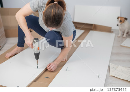 A Caucasian woman uses a screwdriver to tighten a screw while assembling a cabinet. A Caucasian woman uses a screwdriver to tighten a screw while assembling a cabinet. 137873455