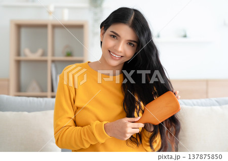 Happy indian woman brushing and detangling hair using wooden brush in living room at home 137875850