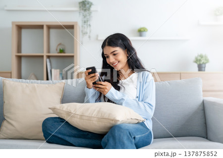 Smiling pretty young Asian woman sitting on cozy couch Smiling pretty young Asian woman sitting on cozy couch 137875852