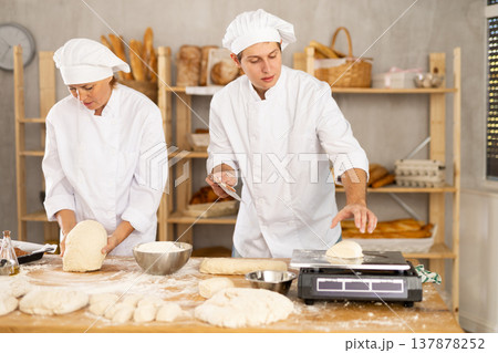 Experienced female adult baker and young male assistant in white uniform kneading dough cutting into pieces and weighing preparing for baking pastries in bakery Experienced female adult baker and young male assistant in white uniform kneading dough cutting into pieces and weighing preparing for baking pastries in bakery 137878252