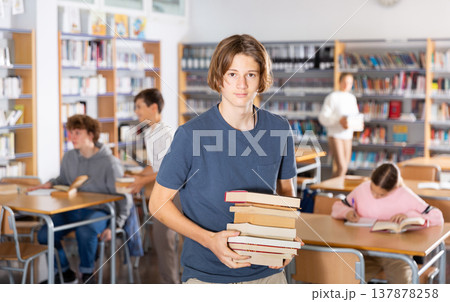Teenager boy standing with a stack of books in library Teenager boy standing with a stack of books in library 137878258