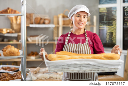 In working room of bakery, there is woman with baguettes in wicker basket in hands In working room of bakery, there is woman with baguettes in wicker basket in hands 137878557
