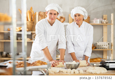 Two bakers, woman and young guy, knead raw dough on table in bakery kitchen 137878582