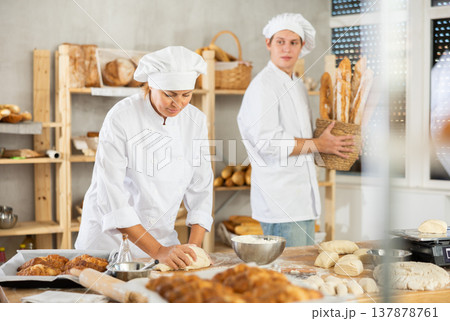 Female baker kneads dough on kitchen counter while young male assistant carries fresh baguettes to bakery display case Female baker kneads dough on kitchen counter while young male assistant carries fresh baguettes to bakery display case 137878761