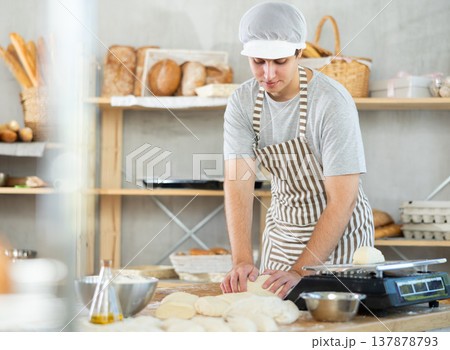 Young positive male baker in apron kneading and forming dough on table, process of preparing of bread in kitchen 137878793