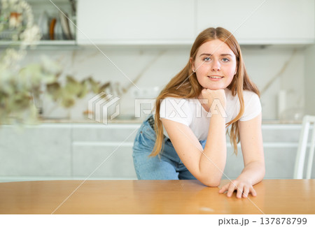 Portrait of positive teenage girl smiling at camera while leaning on countertop in cozy kitchen Portrait of positive teenage girl smiling at camera while leaning on countertop in cozy kitchen 137878799