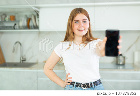 Smiling teenage girl presenting smartphone with blank black screen with copy space in modern cuisine. Free copy space for your design or advertisement Smiling teenage girl presenting smartphone with blank black screen with copy space in modern cuisine. Free copy space for your design or advertisement 137878852