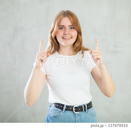 Portrait of cheerful charismatic teen girl in white t-shirt and blue jeans pointing up against gray wall with copy space 137878918