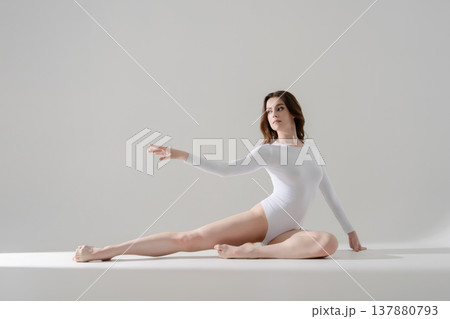 Elegant woman in white leotard posing gracefully on floor in a bright minimalist studio. 137880793