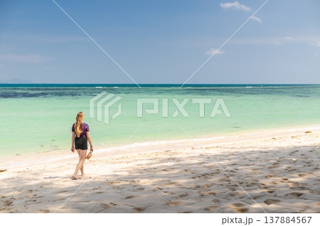 Young woman walking barefoot along sandy beach in Koh Samui, Thailand, holding hat and looking toward turquoise sea under clear blue sky during tropical summer travel Young woman walking barefoot along sandy beach in Koh Samui, Thailand, holding hat and looking toward turquoise sea under clear blue sky during tropical summer travel 137884567