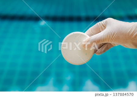 Gloved hand holding round chlorine tablet above turquoise swimming pool water, pool maintenance and sanitation concept with clean background and copy space Gloved hand holding round chlorine tablet above turquoise swimming pool water, pool maintenance and sanitation concept with clean background and copy space 137884570