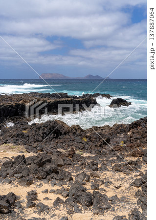 Dramatic seascape of La Graciosa with volcanic rocks in the foreground and islands under a blue sky. 137887064